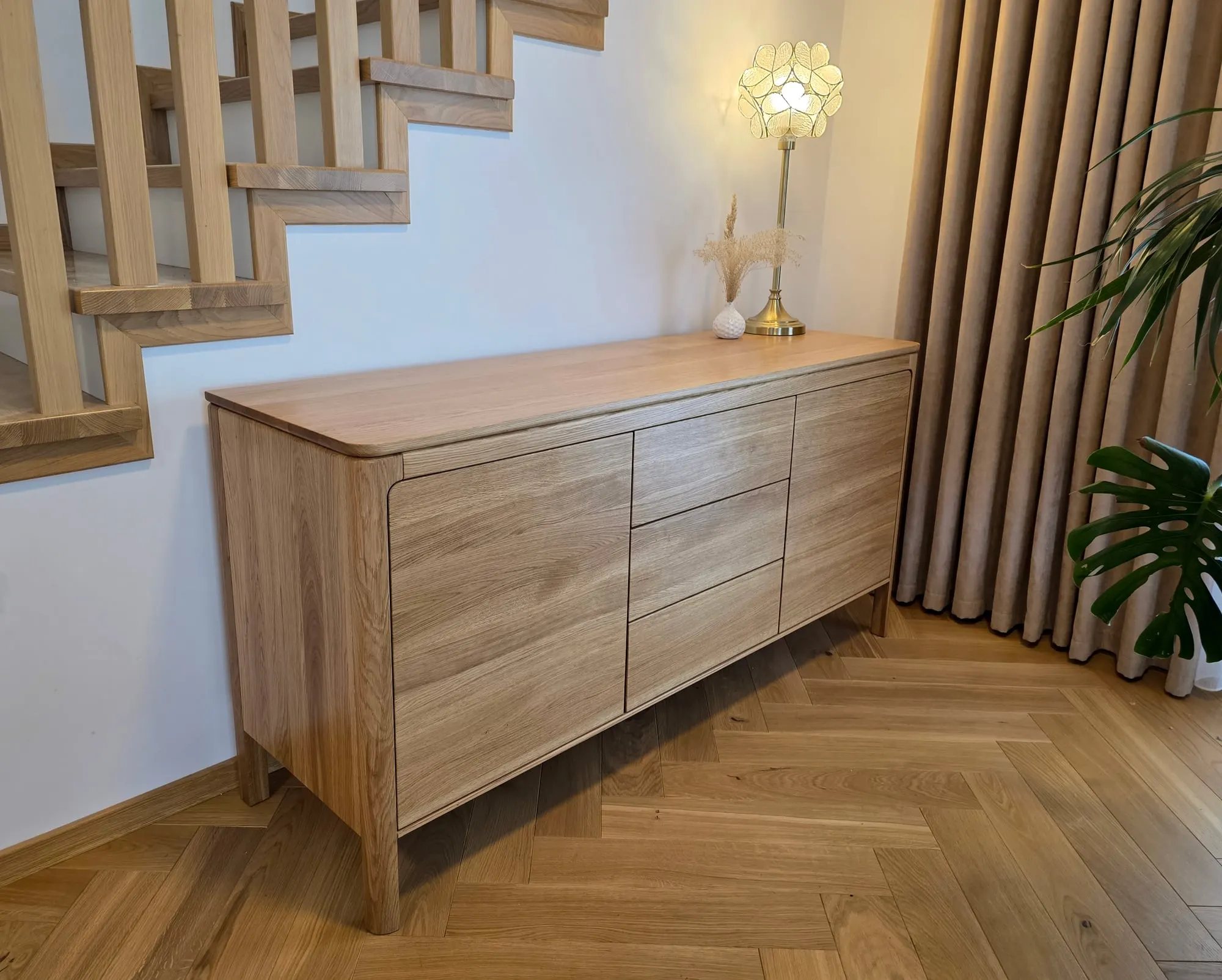 Dębowa komoda o zaokrąglonych narożnikach, ustawiona przy schodach z balustradą.