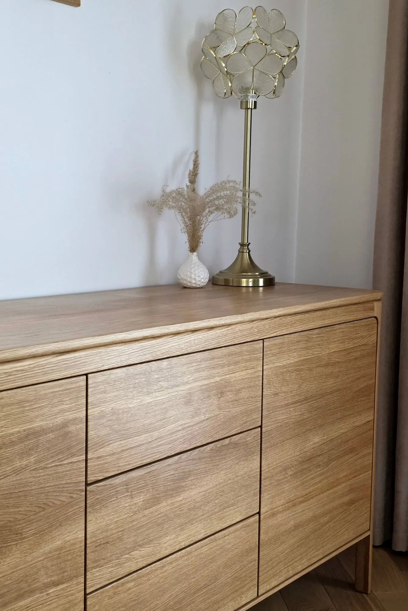 Pionowe ujęcie dębowej komody z lampą na blacie, w sąsiedztwie zasłon.