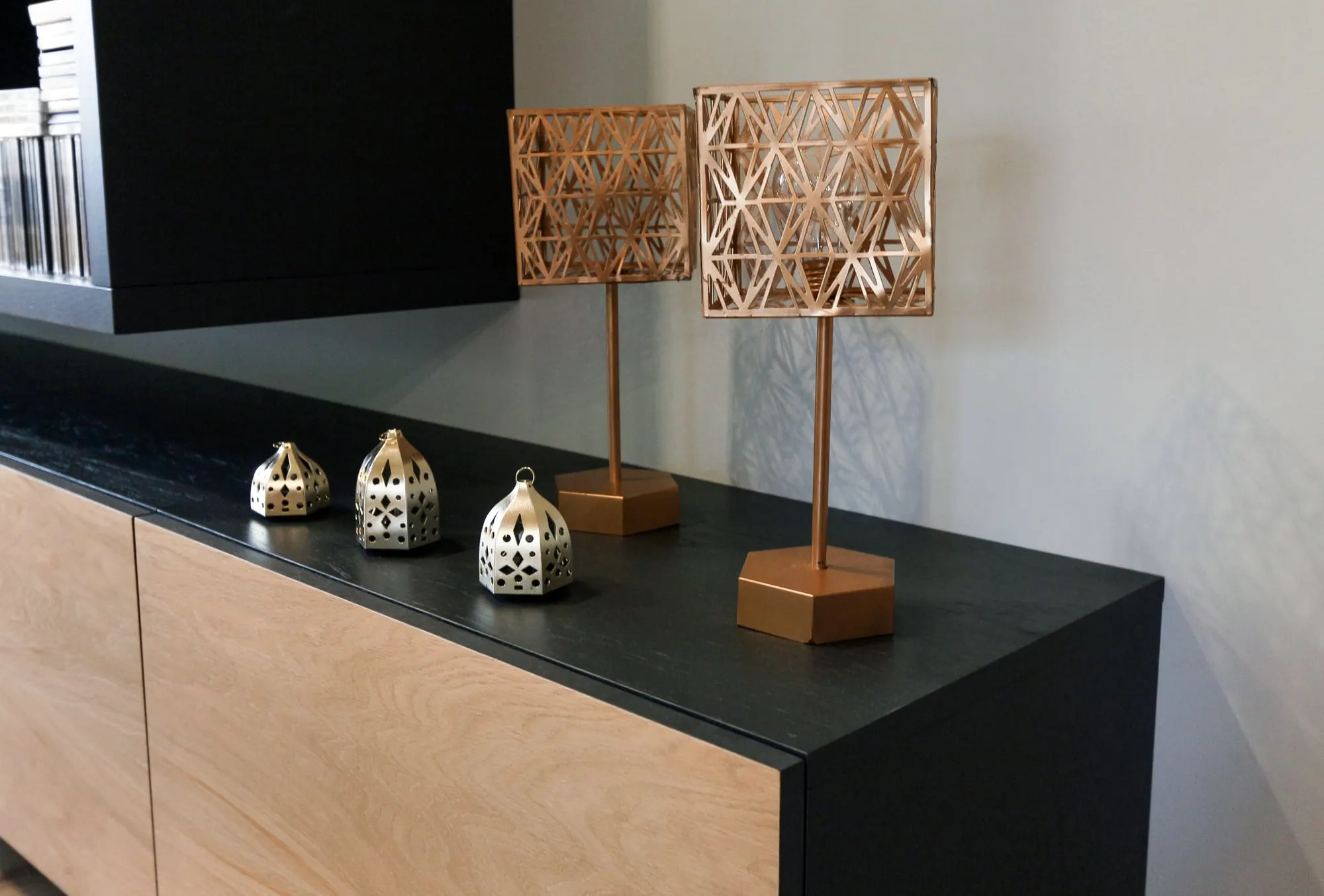 Zbliżenie lampionów i geometrycznych lamp na blacie szafki pod regałem na książki.