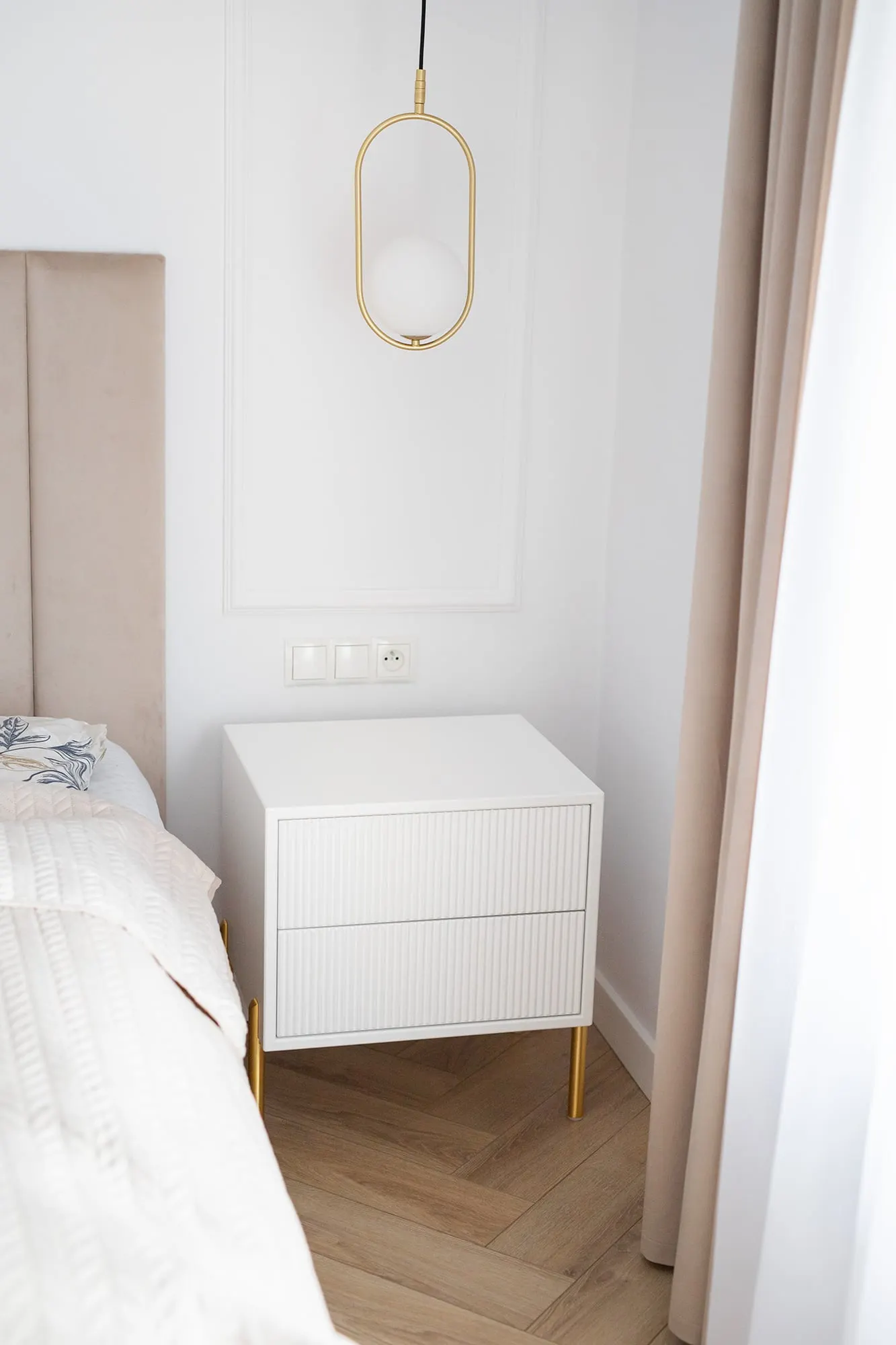 Szafka nocna przy łóżku, nad nią owalna lampa wisząca na złotym stelażu.